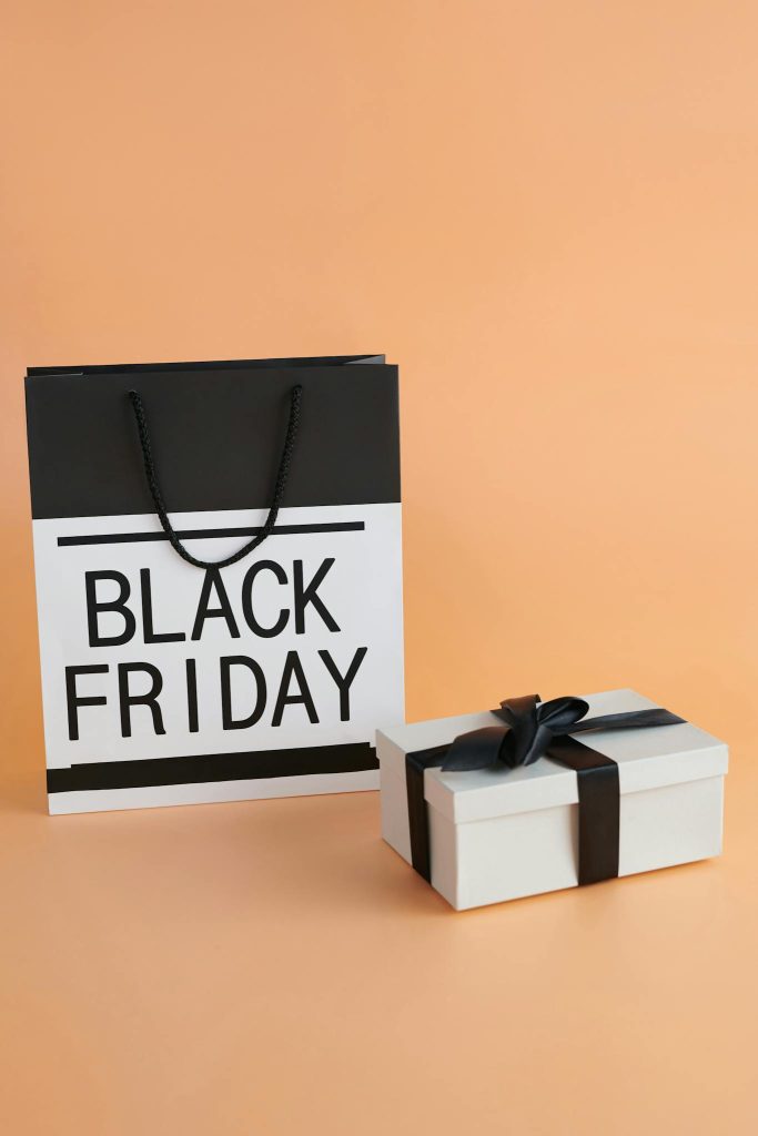 Modern Black Friday shopping bag and gift box with black ribbon against peach background.